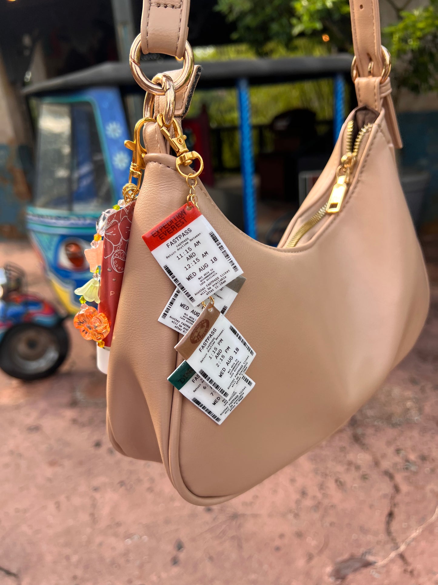 A beige purse with Disney-inspired acrylic FastPass charms clipped to the strap, featuring retro ride tickets like Expedition Everest and Dinosaur in front of the iconic Animal Kingdom bike.