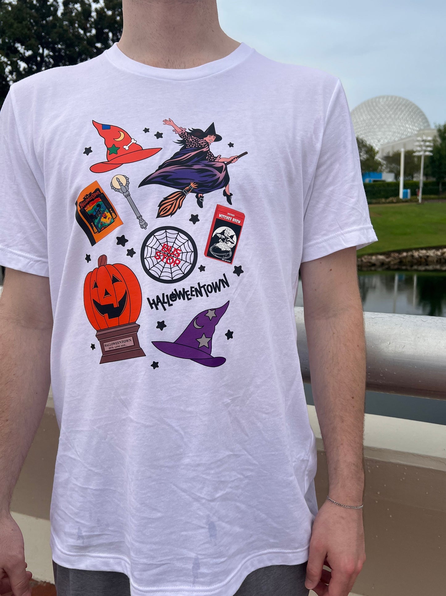 White T-shirt with a collage of Halloweentown-inspired icons including a witch on a broom, pumpkin, spellbook, potion, witch hats, and stars.
