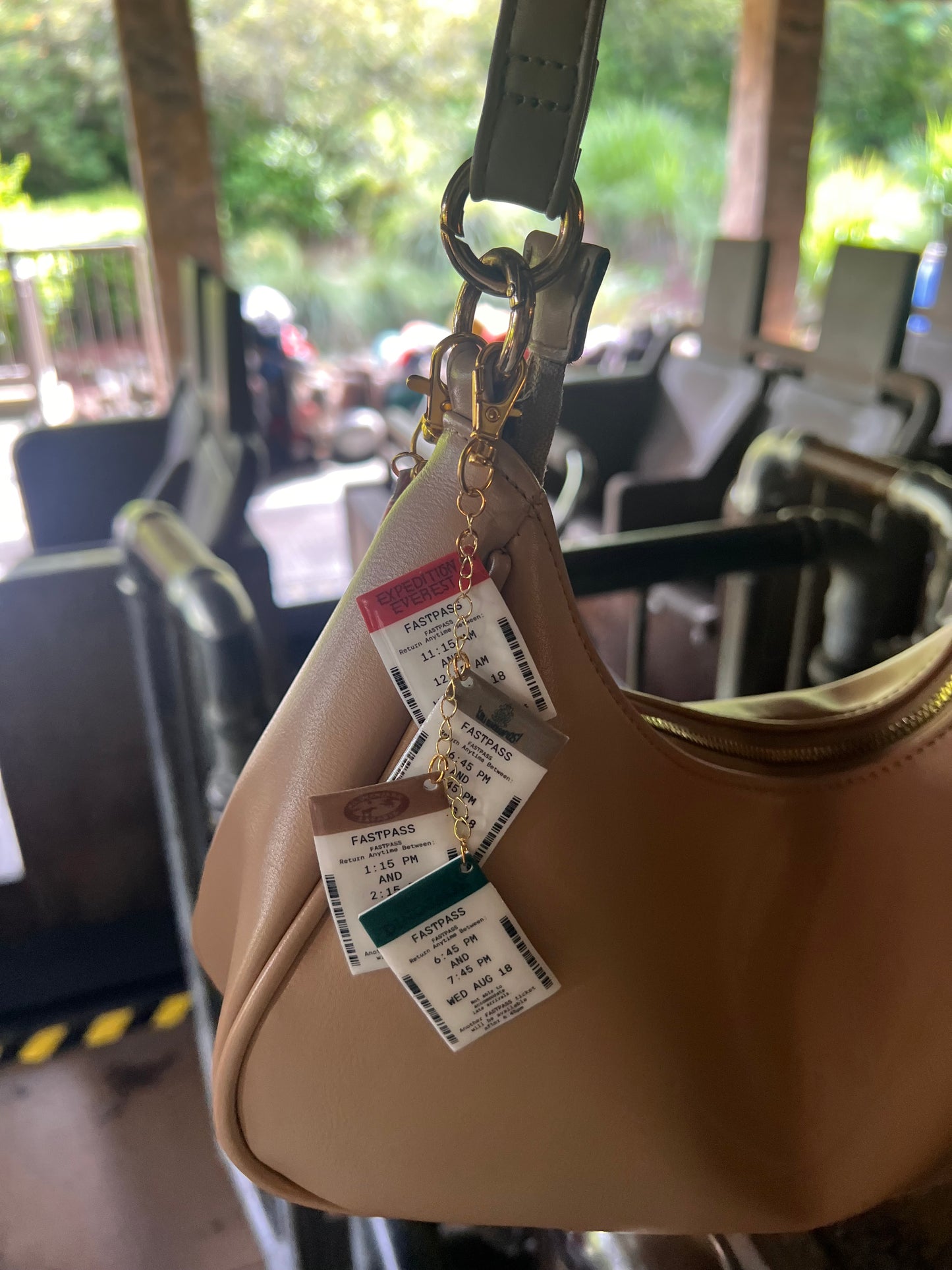 Close-up of Disney FastPass purse charms hanging from a gold keychain on a tan handbag, designed to look like vintage park ride tickets positioned in front of Expedition Everest ride in Animal Kingdom