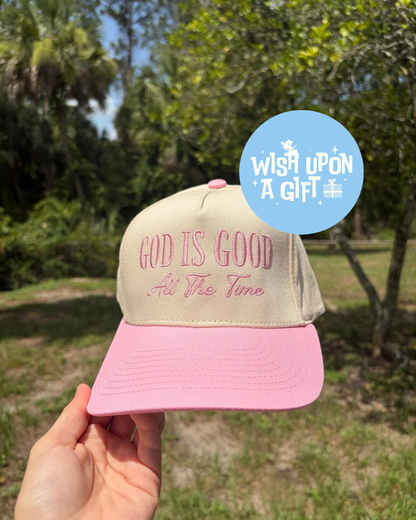 Pink and beige cap with 'God is Good All the Time' text held outdoors.