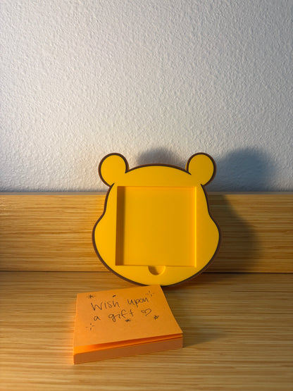 A yellow pooh bear-shaped sticky note holder with a square insert slot, standing on a wooden surface against a textured white wall. An orange sticky note pad with handwritten text lies in front of it.