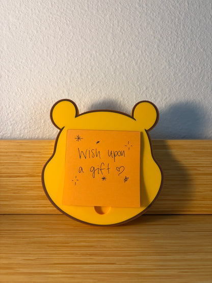 A yellow pooh bear-shaped sticky note holder with a brown outline, holding an orange sticky note that reads 'Wish upon a gift heart' on a wooden surface against a textured white wall.