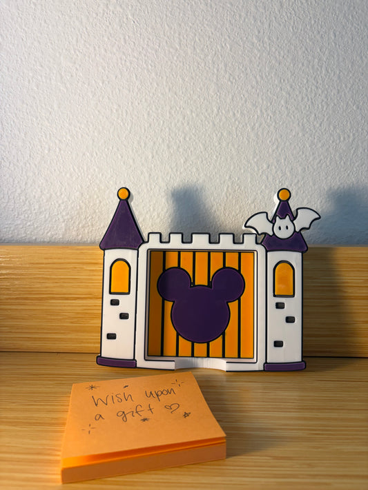 A castle-shaped decorative notepad holder with a purple Mickey Mouse silhouette on the front, orange and purple mickey mouse details, and a small ghost figure on the right tower. It sits on a wooden surface against a white wall, with an orange sticky note in front that reads 'Wish upon a gif.